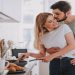 Young bearded man gently kissing his charming girlfriend while she stirring something in pan