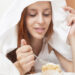 Long-haired woman eating sweet cake under white sheet in bed